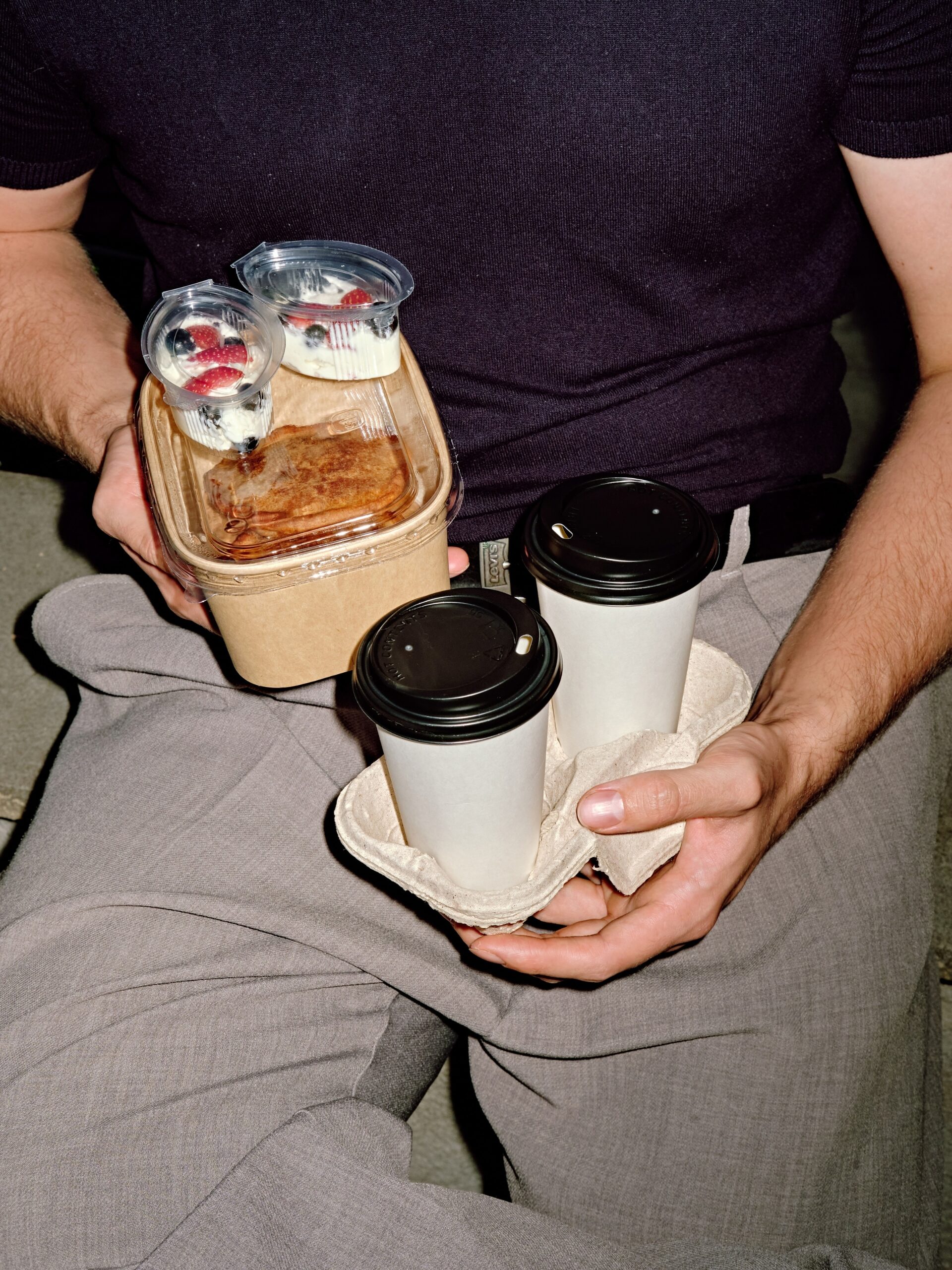 Two takeaway coffees in a carrier and a dessert box on someone's lap