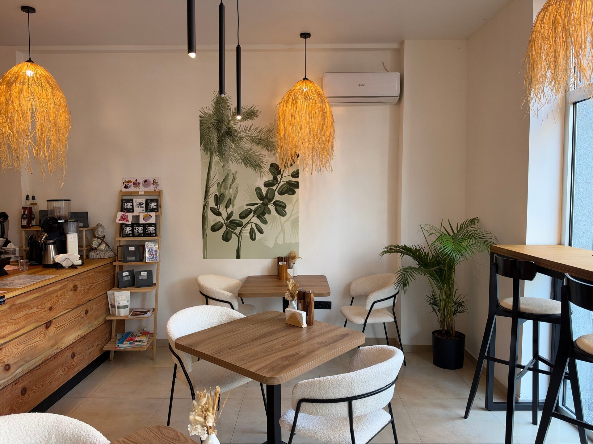 The room seen from the counter: a ladder shelf with drip coffee boxes and bean bags, a small palm mural, wooden tables and a window bar with stools